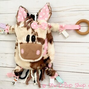 May include: A brown and white cow-print plush toy with pink ears and a pink bow. The toy has a wooden ring attached to it with a pink ribbon. The toy is on a white wooden surface.