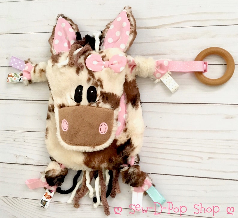 May include: A brown and white cow-print plush toy with pink ears and a pink bow. The toy has a wooden ring attached to it with a pink ribbon. The toy is on a white wooden surface.