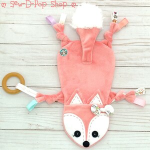 May include: A pink fox-shaped baby toy with a white fur tail, a wooden ring, and various fabric tags. The toy is on a white wooden background.