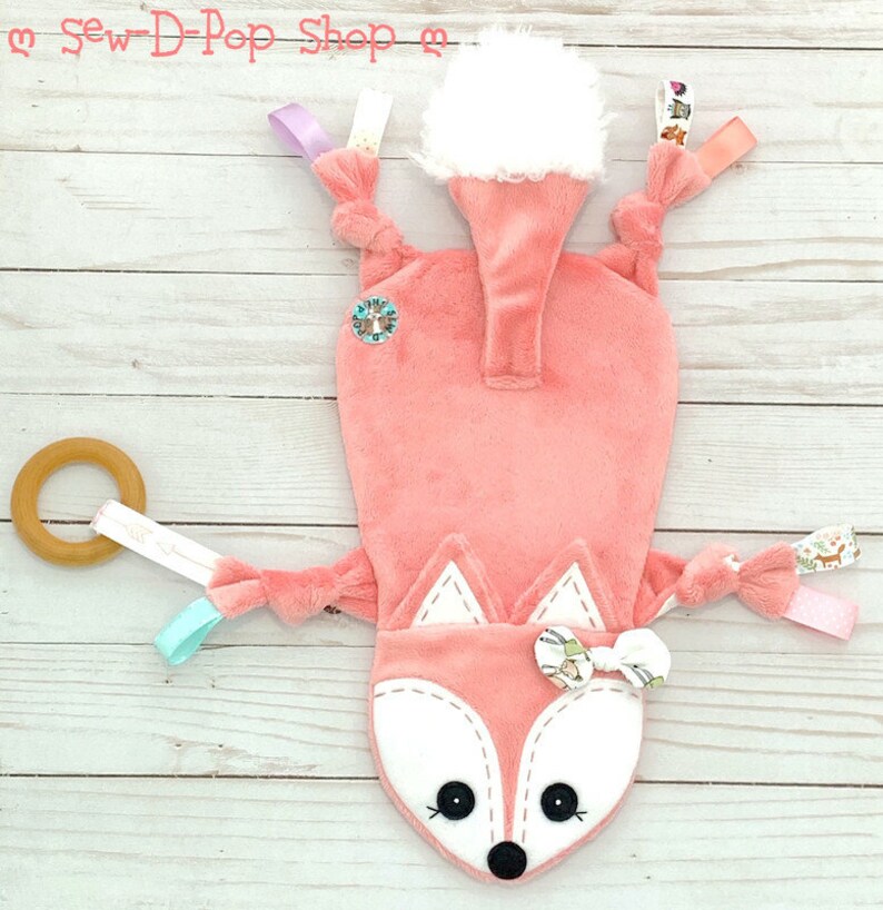 May include: A pink fox-shaped baby toy with a white fur tail, a wooden ring, and various fabric tags. The toy is on a white wooden background.