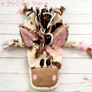 May include: A brown and white spotted plush horse toy with pink ears and a pink bow. The horse has brown yarn hair and black eyes. The toy has a wooden ring attached to a pink ribbon.