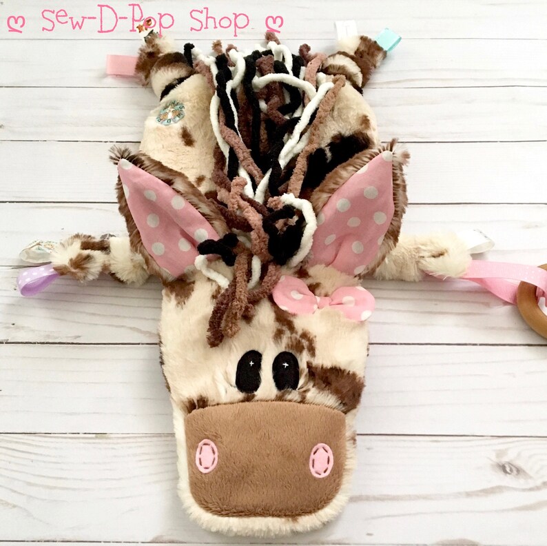May include: A brown and white spotted plush horse toy with pink ears and a pink bow. The horse has brown yarn hair and black eyes. The toy has a wooden ring attached to a pink ribbon.