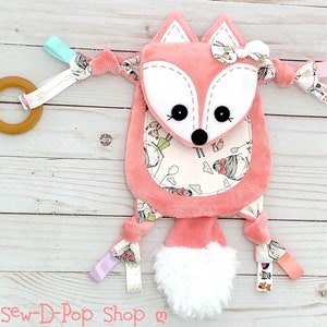 May include: A pink fox-shaped baby toy with a white fluffy tail. The toy has a white and pink patterned fabric with a small fox face on the front. The toy has a wooden ring and a variety of colourful ribbons attached.