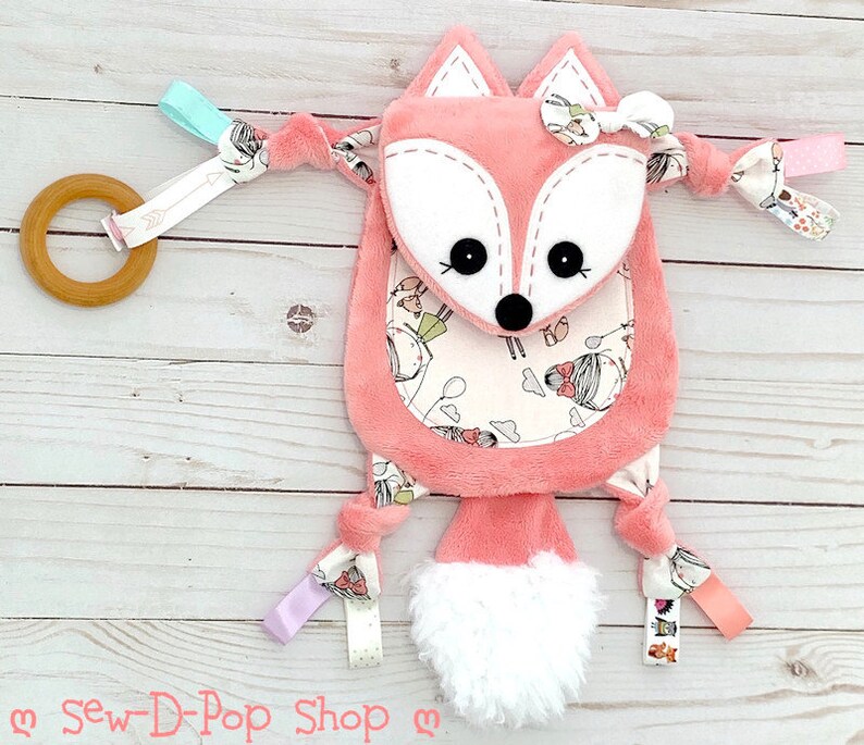 May include: A pink fox-shaped baby toy with a white fluffy tail. The toy has a white and pink patterned fabric with a small fox face on the front. The toy has a wooden ring and a variety of colourful ribbons attached.