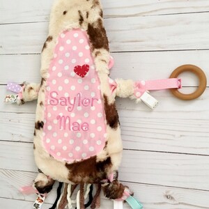 May include: A pink and brown cow-print baby toy with a pink polka dot fabric panel. The panel has the name "Saylor Mae" embroidered on it in white thread. The toy has a wooden ring attached to a pink ribbon and several fabric tags.