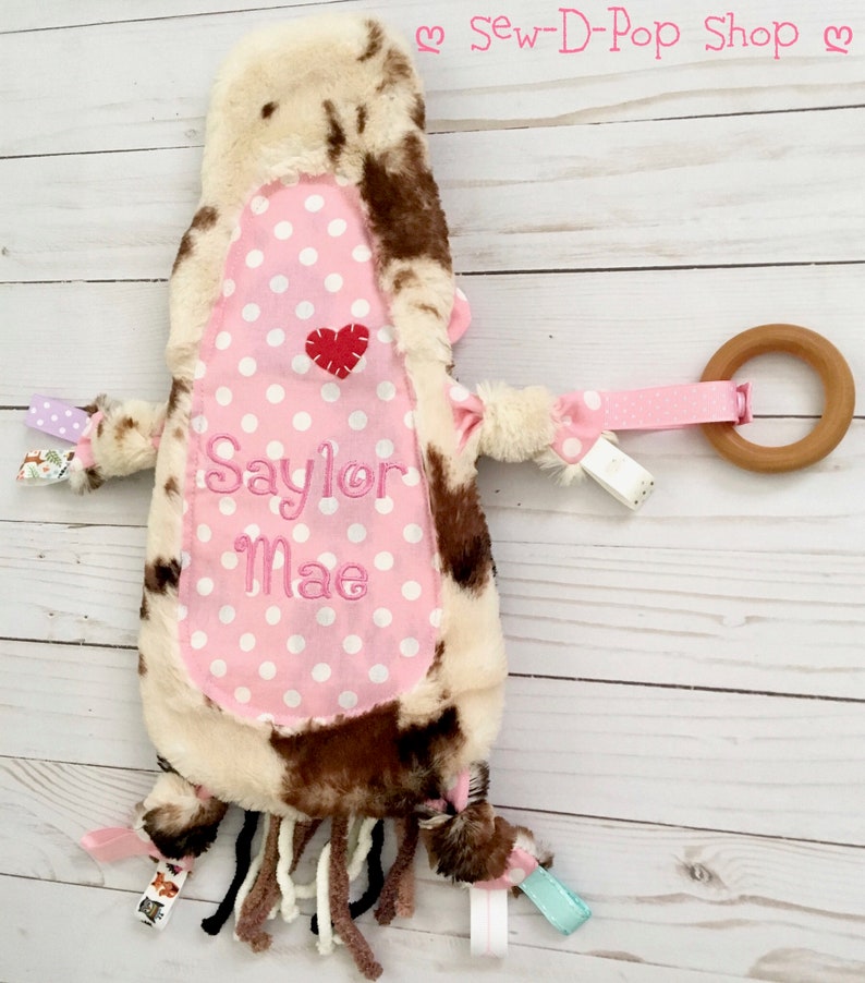 May include: A pink and brown cow-print baby toy with a pink polka dot fabric panel. The panel has the name "Saylor Mae" embroidered on it in white thread. The toy has a wooden ring attached to a pink ribbon and several fabric tags.