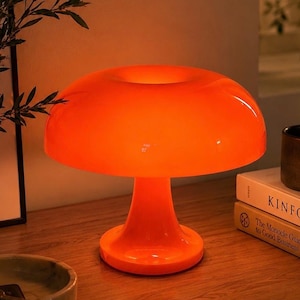 Könnte beinhalten: Eine leuchtend orangefarbene, pilzförmige Tischleuchte mit glatter, glänzender Oberfläche und einem breiten, abgerundeten Schirm, der ein warmes, leuchtendes Licht abgibt. Die Lampe hat einen zentralen Sockel, der sich nach unten hin verbreitert.