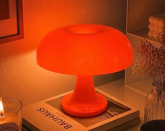 Puede incluir: Una lámpara de mesa de color naranja brillante con forma de seta, acabado liso y brillante, y una pantalla de cúpula ancha y redondeada. La lámpara emite un resplandor cálido y vibrante sobre la superficie de un libro titulado BAUHAUS debajo.
