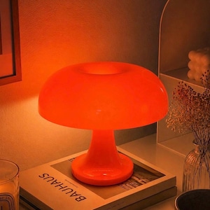Op de afbeelding: Een oranje paddenstoelvormige tafellamp. De lamp heeft een afgeronde kap en een voetstuk. Hij staat op een stapel boeken met het woord "BAUHAUS" zichtbaar. De lamp geeft een oranje gloed.