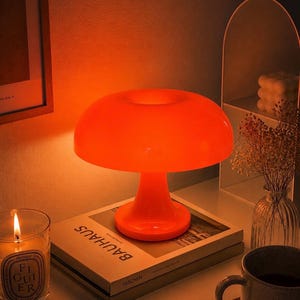 Könnte beinhalten: Eine orangefarbene Tischlampe in Pilzform steht auf einem Stapel Bücher mit den Titeln "Bauhaus" und "Taschen". Eine brennende Kerze und eine Tasse befinden sich ebenfalls auf dem Tisch. Die Lampe strahlt ein warmes Licht aus.