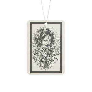 May include: A rectangular hanging air freshener featuring a black and white ink-style illustration of a person with horns holding a traditional mask. The design includes intricate floral details, swirling smoke elements, and a thin white string.