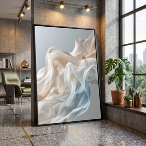 May include: A large framed art print with an abstract design of flowing fabric in white, beige, and light blue hues. The artwork is displayed in a modern interior setting, leaning against a wall.