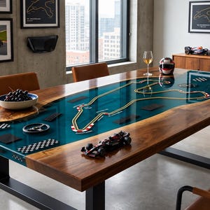 Puede incluir: Una mesa de comedor rectangular con borde de madera natural y una incrustación central de resina azul que representa un circuito de Fórmula 1. La mesa incluye elementos de coches de carreras en miniatura, un volante y patas de metal negro.