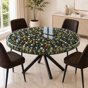 Può includere: Un tavolo da pranzo rotondo con un piano in vetro riempito di numerose piccole sfere di vetro multicolori nei toni del blu, del verde e dell'ambra. Il tavolo presenta una base in metallo nero con gambe angolate e quattro sedie imbottite marroni.