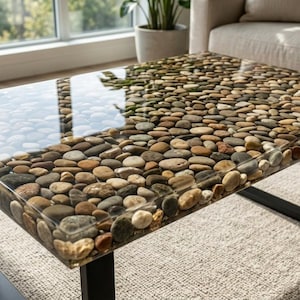 Peut inclure: Une table basse rectangulaire dotée d'un plateau en résine transparente incrusté d'une couche dense de galets de rivière lisses aux tons gris, marron et beige. La table est soutenue par un cadre en métal noir avec quatre pieds.