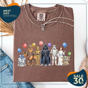 a brown t-shirt with a graphic design of various Star Wars characters, including Chewbacca, Darth Vader, and the Child, along with balloons. The t-shirt is displayed on a white background, and there are jeans and a sweater visible in the background.