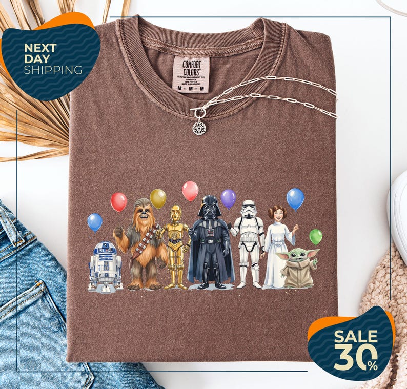 a brown t-shirt with a graphic design of various Star Wars characters, including Chewbacca, Darth Vader, and the Child, along with balloons. The t-shirt is displayed on a white background, and there are jeans and a sweater visible in the background.