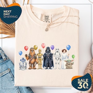 a t-shirt with a graphic design of various Star Wars characters, including Chewbacca, Darth Vader, and the Yoda from the Star Wars franchise, along with balloons. The t-shirt is displayed on a white background, and there are jeans visible in the background.