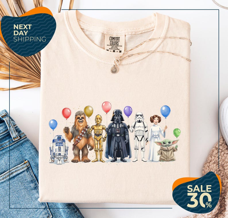 a t-shirt with a graphic design of various Star Wars characters, including Chewbacca, Darth Vader, and the Yoda from the Star Wars franchise, along with balloons. The t-shirt is displayed on a white background, and there are jeans visible in the background.