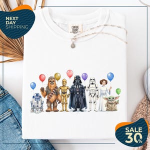 a white t-shirt with a graphic design of various characters from popular media, including Chewbacca, Darth Vader, Star Wars, and Baby Yoda, holding colorful balloons.