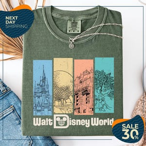 a green t-shirt with a graphic design of Disney World landmarks, including a castle, roller coaster, and other iconic structures. The t-shirt is displayed on a white background, and there are two sale tags visible, one for the t-shirt and one for a pair of jeans.