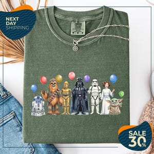 a green t-shirt with a graphic design of various Star Wars characters, including Chewbacca, Darth Vader, and the Child, along with balloons.