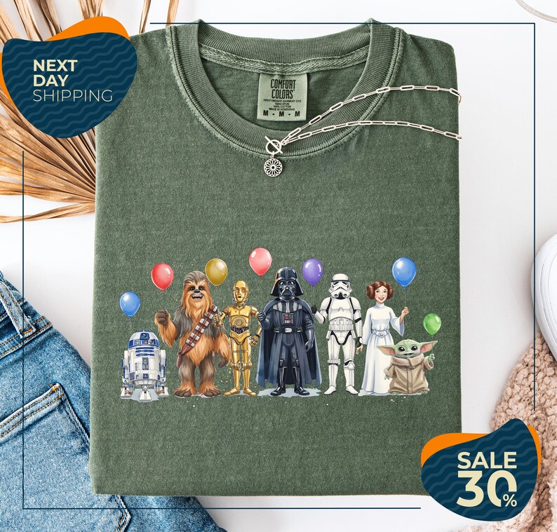 a green t-shirt with a graphic design of various Star Wars characters, including Chewbacca, Darth Vader, and the Child, along with balloons.