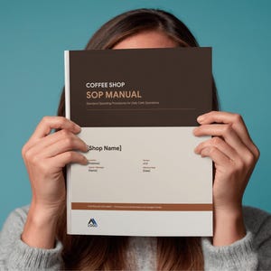 May include: A person holding a coffee shop SOP manual. The cover is brown and white with the title "COFFEE SHOP SOP MANUAL" and "Standard Operating Procedures for Daily Cafe Operations". The manual has a blue background.