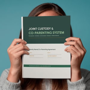 May include: A white and green booklet titled "JOINT CUSTODY & CO-PARENTING SYSTEM" is held in front of a person's face. The booklet includes sections for parenting plans, schedules, communication, expenses, and children's records.