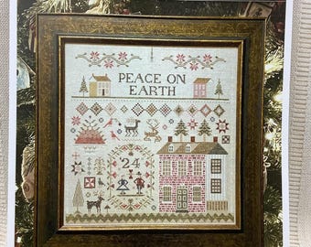 May include: A framed cross-stitch sampler titled PEACE ON EARTH BY SHAKESPEARE'S PEDDLER. The design features a brick house, a couple, reindeer, trees, and geometric patterns in red, green, and brown thread on a light fabric background.