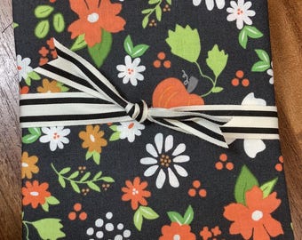 May include: A rectangular book or journal cover featuring a black fabric background with a repeating pattern of orange and white flowers, green leaves, and small orange pumpkins, tied with a black and white striped fabric ribbon.