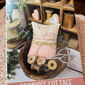 May include: A book cover with the title "Abide In My Love" and the author's name, Lindsey Weight. The cover features a pink cross-stitched pillow with the title embroidered on it, along with wooden spools, scissors, and the text "Primrose Cottage".