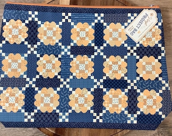 May include: A rectangular fabric project bag featuring a repeating quilt-style pattern of peach-colored floral blocks on a dark navy blue background. An orange zipper runs along the top edge, and a white tag reads Vinyl PROJECT BAG.