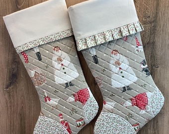 May include: Two quilted Christmas stockings featuring a beige background with a repeating pattern of Santa Claus, gingerbread houses, and geese. The stockings have a solid cream cuff and a patterned toe section with small red berries.