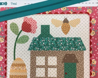 May include: A fabric quilt block featuring a house with a green door, a brown chimney, and white windows. A large pink flower and a yellow bee are appliqued on a cream background. The block is bordered by pink floral fabric and a teal zipper.