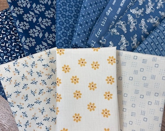 May include: A collection of folded fabric squares featuring various patterns in shades of navy blue, white, and yellow. Designs include floral prints, geometric shapes, and text that reads, "see the good in this day, this is one moment in time."