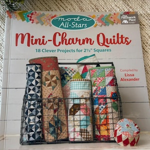 May include: A book titled "Mini-Charm Quilts" with the text "18 Clever Projects for 2 1/2" Squares." The book is next to a wire basket filled with rolled fabric bundles. The book cover has a colorful patchwork design.