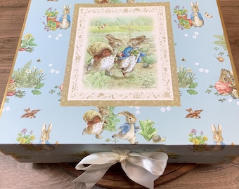 May include: A square light blue gift box featuring a repeating Peter Rabbit pattern and a central illustration of two rabbits in a garden. The box is tied with a white satin ribbon bow at the front and includes the text Peter Rabbit.