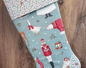 May include: A light blue Christmas stocking featuring a repeating pattern of a nutcracker, a goose with a red bow, a gingerbread house, a drum, and Santa Claus. The cuff and toe are accented with a white fabric featuring green holly leaves and red berries.