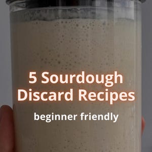 Puede incluir: Un tarro de vidrio transparente con tapa metálica, lleno de una mezcla beige y burbujeante. El texto "5 Sourdough Discard Recipes" y "beginner friendly" se muestra en el centro del tarro. El tarro es sostenido por una mano.