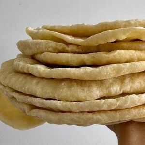 May include: A stack of round, beige, flatbreads with a slightly uneven, textured surface. The edges of the circular breads are soft and rounded, showing a light golden-brown colour. The stack consists of multiple layers of thin, cooked dough.
