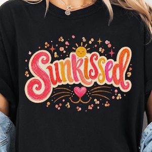 以下が含まれることがあります： 黒いTシャツに、カラフルで遊び心のあるフォントで「Sunkissed」の文字。デザインには、笑顔の太陽、ハート、装飾的な要素が含まれています。カジュアルな普段着アイテムです。