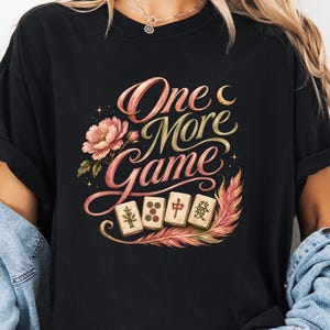 Puede incluir: Una camiseta negra con un estampado gráfico que incluye el texto "One More Game" en letra cursiva elegante. El diseño presenta una flor de peonía rosa, cuatro fichas de mahjong, una luna creciente, estrellas pequeñas y una pluma colorida.