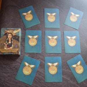 Może przedstawiać: Talia kart "Angels and Ancestors Oracle Cards" z pudełkiem przedstawiającym kobietę ze skrzydłami anioła. Karty są w kolorze morskim ze złotym i białym wzorem. Karty są rozłożone na ciemnej powierzchni.