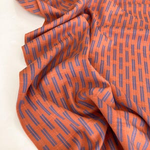 May include: A piece of fabric featuring a vibrant orange base colour with a repeating pattern of vertical, dashed blue lines. The material displays a textured, woven appearance with folds that create natural shadows across the surface.