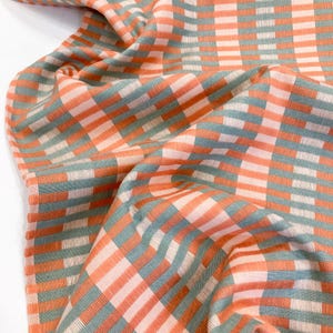 May include: A woven fabric featuring a repeating geometric pattern of vertical and horizontal stripes in shades of coral orange, muted sage green, and soft cream. The textile displays a textured, grid-like weave with visible thread intersections.