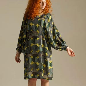 Könnte beinhalten: Ein knielanges Kleid mit einem sich wiederholenden Muster aus dunkelgrünen und grauen Kreisen auf gelbem Grund. Das Kleidungsstück hat einen Rundhalsausschnitt und lange, voluminöse Ärmel, die an den Handgelenken mit einem dezenten Bündchen abschließen.