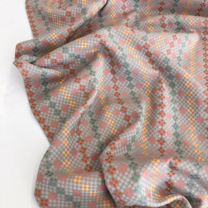 May include: A woven fabric featuring a dusty rose background with a repeating geometric diamond pattern in shades of teal, mustard yellow, and terracotta. The textile displays a textured, intricate weave with soft folds and draped fabric sections.