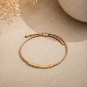 May include: A bracelet featuring a thin, tan-colored cord and a smooth, curved gold-toned metal bar. The design includes a sliding gold-toned metal clasp mechanism and small gold-toned metal end caps on the cord, creating a simple, adjustable piece.