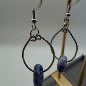 May include: A pair of silver-toned teardrop earrings. Each earring features a blue and white speckled bead suspended within the teardrop shape. The earrings have a simple, elegant design.